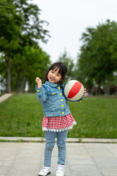 Cute little Asian girl in a denim jacket standing on a modern outdoor pavement, holding a colorful toy ball and smiling at the camera, happy childhood play concept.