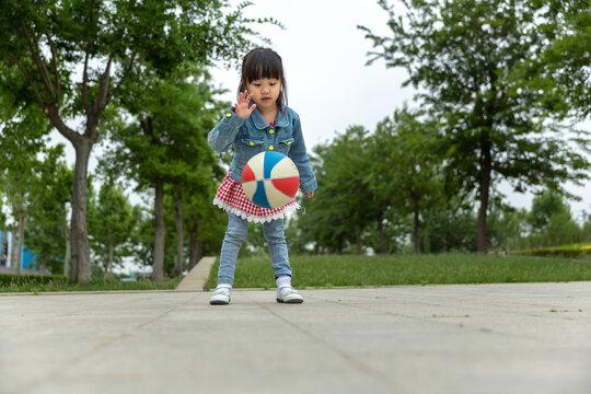 Cute little Asian girl focusing on bouncing a colorful toy ball on a park paved ground, practicing gross motor skills and coordination, outdoor early education.
