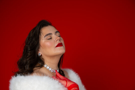 Photo of young adult white woman with closed eyes, diamond necklace, red lips, tulle gloves, downy fur cape on scarlet background. Concept of March 8, fashion, diverse beauty, luxury, diversity, style