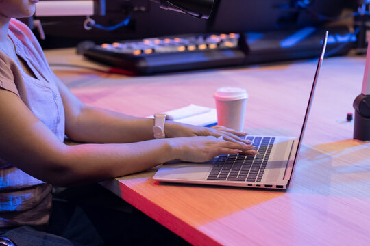 Adult African American woman typing on laptop at studio desk with mic, mixer, light tee, smartwatch