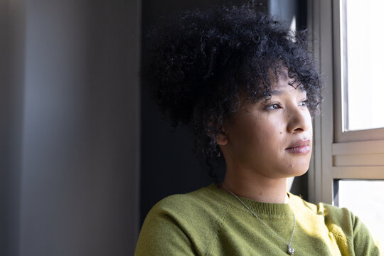 Woman sitting by window, gazing out with pendant necklace, small earrings and green sweater
