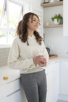 Teen African American female leaning on kitchen counter, holding glass bottle, wearing cream hoodie