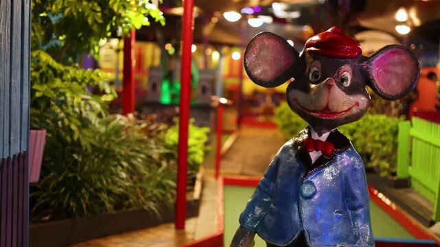 A happy mouse statue wearing a blue suit and red hat stands cheerfully in front of a vibrant miniature golf course at night, colorful lights illuminate the fun and lively atmosphere.