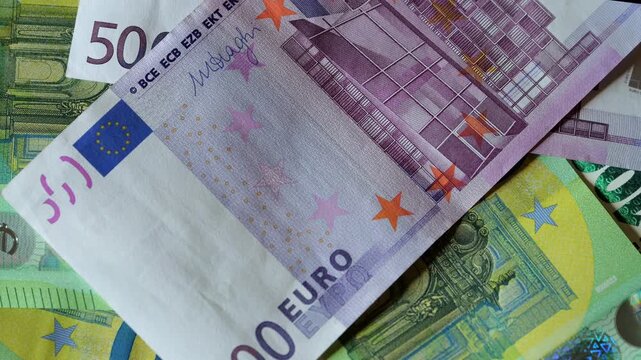 Close-up of fifty euro banknote. Architectural detail visible on paper. Hundred euro note layered below. Green and purple tones dominate the scene. Star patterns and currency text clearly shown