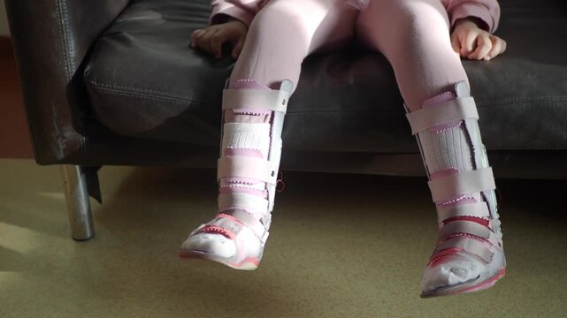 Little girl with cerebral palsy sitting on sofa wearing orthopedic leg braces for physical therapy and walking assistance