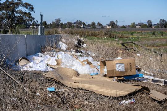 Illegally dumped waste, cardboard boxes, plastic packaging, and debris on a vacant peri-urban road reserve bordering new residential housing estates. Truganina, Melbourne, Victoria, Australia