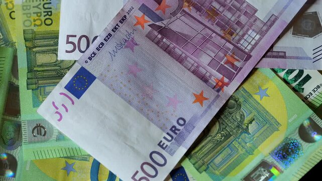 Stack of euro banknotes, including 500 euro notes. Currency pile shows various denominations. Notes feature architectural designs and stars. Background contains blurred green and yellow notes