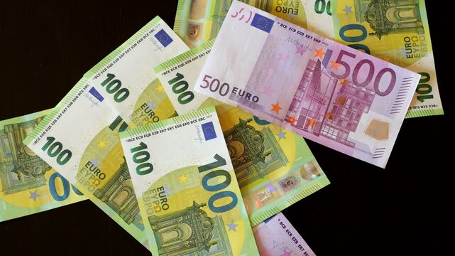 Euro banknotes scatter across dark surface. One hundred and five hundred euro bills visible. Notes fall slowly, motion captured in detail. Shiny paper currency spread out naturally