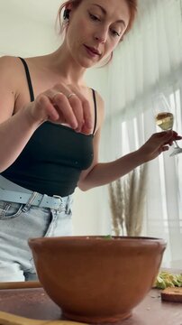 Mid adult millennial woman preparing a healthy green salad at home while drinking wine, dancing and singing, enjoying a relaxed and playful moment during everyday cooking.