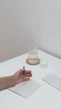 Hands Writing in Notebook with Pen on Desk Beside Drinking Glass and Computer Keyboard