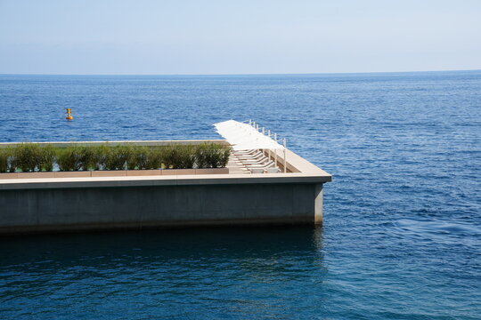 Modern seaside deck with a row of white sun loungers and umbrellas overlooking a vast blue sea under a pale, cloudless sky.