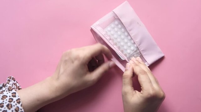 Woman keeping birth control pills in zipper pouch, person holding contraception pills in blister pack, top view of female hands with contraceptive tablets