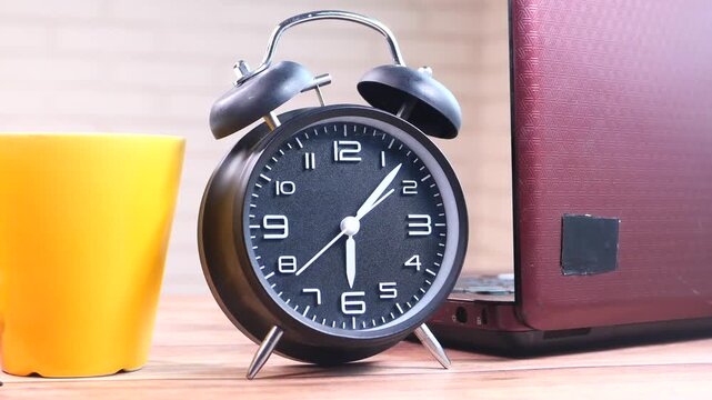 Black alarm clock standing on wooden table next to laptop and yellow mug
