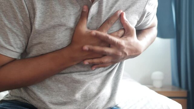 Unrecognizable man suffering from sudden heart attack or chest pain while sitting on bed at home