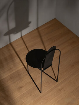 A minimalist black chair sits on a warm wooden floor, illuminated by soft natural light. Long shadows from the chair and nearby objects add depth to the simple interior.