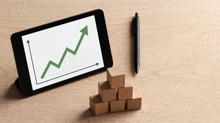 Fototapeta premium Business Growth and Financial Success Concept with Digital Tablet Showing Rising Green Stock Market Graph Next to Pyramid of Wooden Blocks and a Professional Pen on a Light Wood Desk Surface