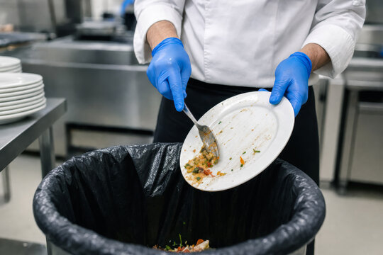 Faceless chef wearing blue gloves scrapes leftover food from a plate into a trash bin in a commercial kitchen, illustrating restaurant cleanup, food waste disposal, hygiene, and sanitation.