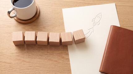 Fototapeta premium Conceptual business growth image with wooden blocks leading to a rocket launch symbol on paper with a cup of coffee and leather notebook on a desk, symbolizing startup success and planning.