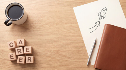 Fototapeta premium Business career growth and startup concept featuring wooden blocks spelling CAREER with a hand-drawn rocket on paper, leather notebook, pen, and a cup of black coffee on a light wooden desk surface.