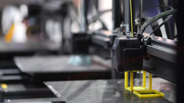 3D printer extrudes yellow plastic. Machine prints on metal surface. Focused shot of printing head in motion. Background blurred with industrial equipment