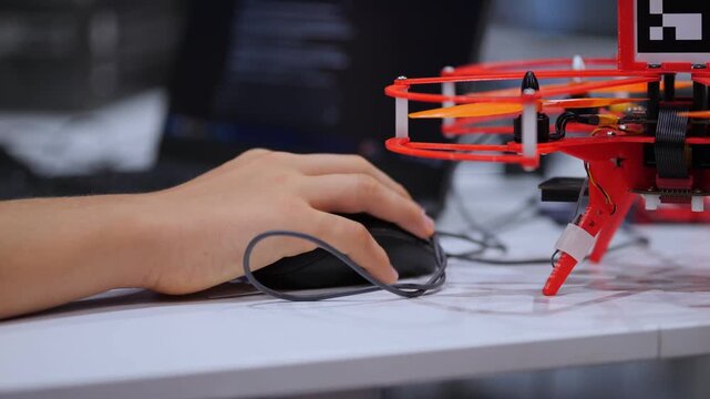 Hand controls computer mouse near red drone model on desk. Drone stands beside mouse and blue pen. Background shows blurred laptop screen with code. White desk surface under objects