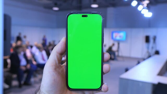 Hand holds phone with green screen. Background shows blurred indoor event space. Audience seated in rows. Lights and stage visible. Phone centered in frame. Smooth camera focus. Realistic lighting
