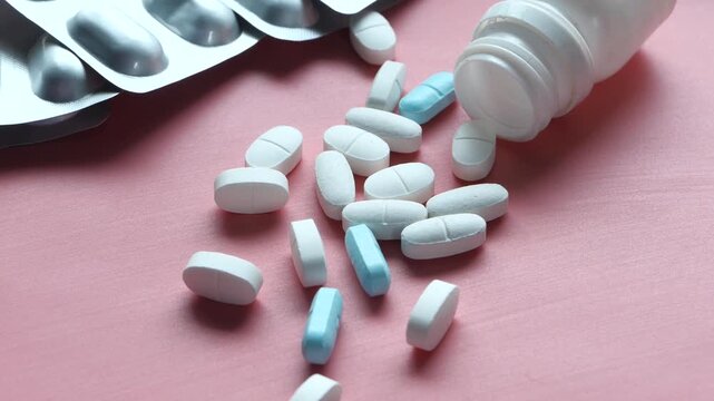 Close-up slow motion video of white and blue pills spilling out of bottle on pink background, medical supplies for health care and treatment