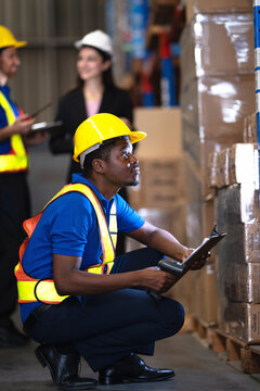 Warehouse worker crouches at the racking to check a barcode reader against the cartons in front of him as a manager reviews a tablet in the aisle just behind him