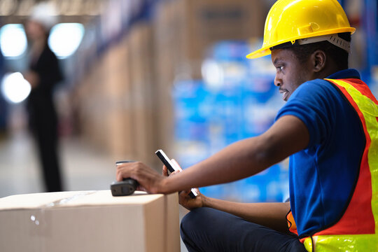 Warehouse employee in a blue polo and orange reflective vest points a barcode reader at a shipping box, a silhouetted co-worker walking the aisle far behind in a distribution centre