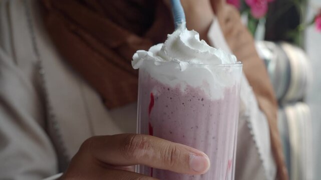 Close up of person in hijab drinking strawberry milkshake topped with whipped cream through straw