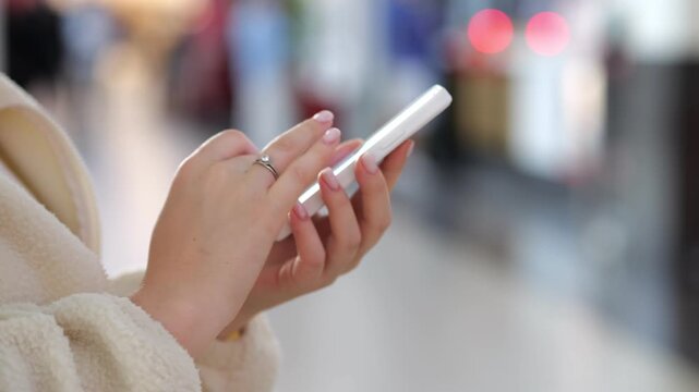 Hands hold smartphone in public space. Background blurred, focus on device and fingers. Person wears light-colored coat. Ring visible on finger. Urban setting with soft lighting