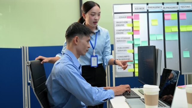 Two software developers collaborate on coding tasks at a computer in a modern tech office with a Kanban board. IT, business