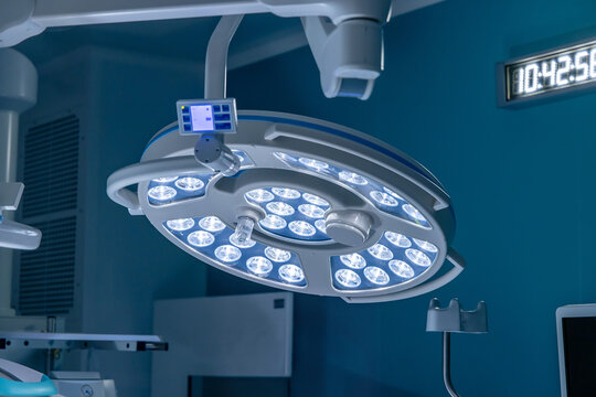 A round LED surgical lamp with a control panel hangs in a modern operating room. A digital clock glows on the wall behind it. The operating theater is prepared for the next surgery