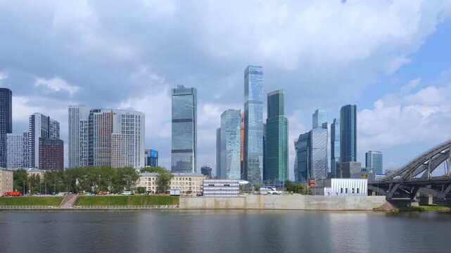 Timelapse of the Moscow City skyline seen across the Moskva River. A steel arch bridge spans the water on the right. Cumulus clouds sail over the glass towers and the granite embankment below.