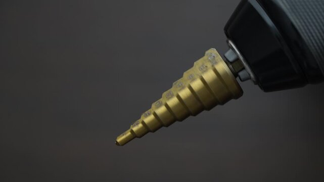 Conical step drill clamped in electric screwdriver rotates, close up. Step conical drill bit for drilling holes with different diameters on the wooden background. Hss step drill bit
