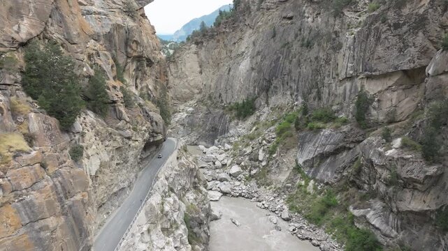 Aerial video of a white vehicle on a gorge road passing beneath towering orange-brown rock walls with patches of green vegetation clinging to the cliff face in Spiti Valley Himachal Pradesh India