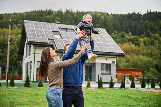 Parents lifting happy child in air on lawn near modern house with solar panels. Photovoltaic rooftop system, renewable energy concept, eco friendly family lifestyle outdoors