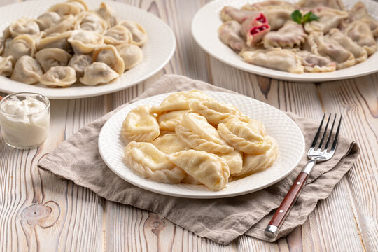 Three white plates with assorted cooked vareniki with different fillings and meat dumplings on rustic wooden background