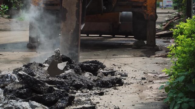 Static tripod shot of a powerful hydraulic breaker attachment breaking asphalt pavement during road construction. Heavy machinery fractures the roadway, creating rubble, debris, and dust at worksite.