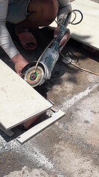 A young Asian man operates a cutting tool on a construction site. He is focused on cutting large tiles on the ground.