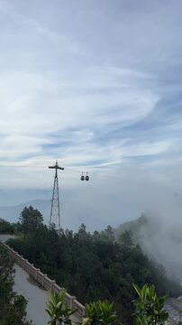 A cable car system travels above a mountainous landscape. The scene includes a power pole and lush greenery under a cloudy sky.
