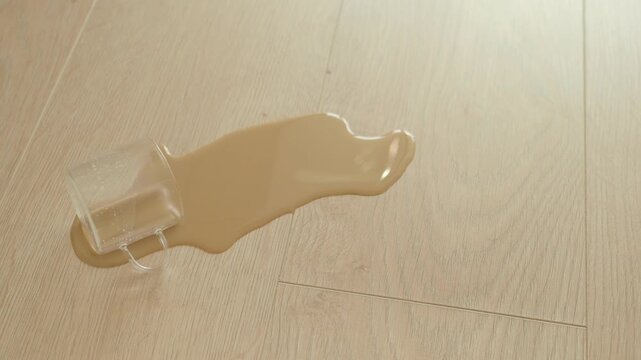 A close up view of an overturned clear plastic cup with light brown liquid spilling out onto a light wood floor