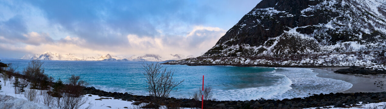Panoramic winter landscape of a coastal beach in Lofoten Islands, Norway. Majestic snow covered mountains over turquoise Arctic ocean waves. Beautiful wild nature background of Scandinavian road trip.