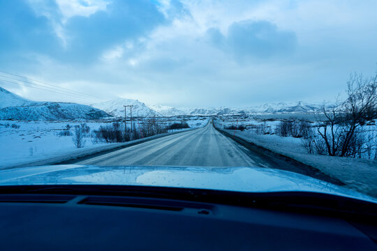 POV driving on an empty asphalt road during a winter road trip in Lofoten Islands, Norway. View from inside the car dashboard looking out at spectacular snow covered mountains and Arctic nature landsc