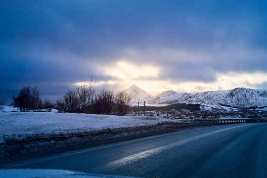 Scenic winter highway road trip in Lofoten Islands, Norway. Driving road with majestic snow covered mountain peak illuminated by sun breaking through dramatic clouds. Beautiful Arctic nature landscape
