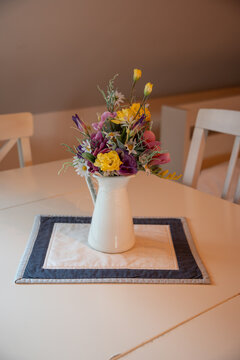 A close-up of a white vase with plastic flowers on a white table with chairs.