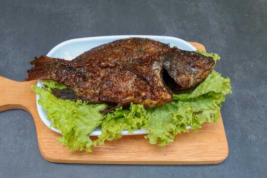 Traditional grilled fish glazed with sweet soy sauce, served on a wooden board with fresh green lettuce. Grilled fish with a dark caramelized exterior, smoky texture