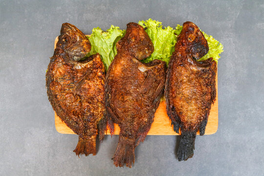 Traditional grilled fish glazed with sweet soy sauce, served on a wooden board with fresh green lettuce. Three whole grilled fish with a dark caramelized exterior, smoky texture