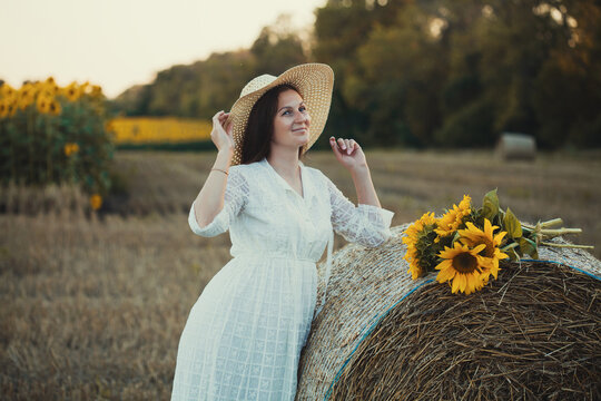 woman in a white dress holding a bouquet of wild flowers with sunflowers in a mowed field with straw bales