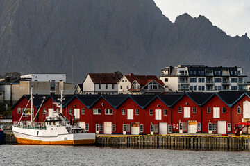 Traditional red wooden fishing huts and white boat in a scenic nordic harbor beneath a steep dark mountain wall with copy space © Quality by Simon